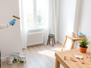 Persoană vopsind perete alb într-o cameră cu parchet şi perdele